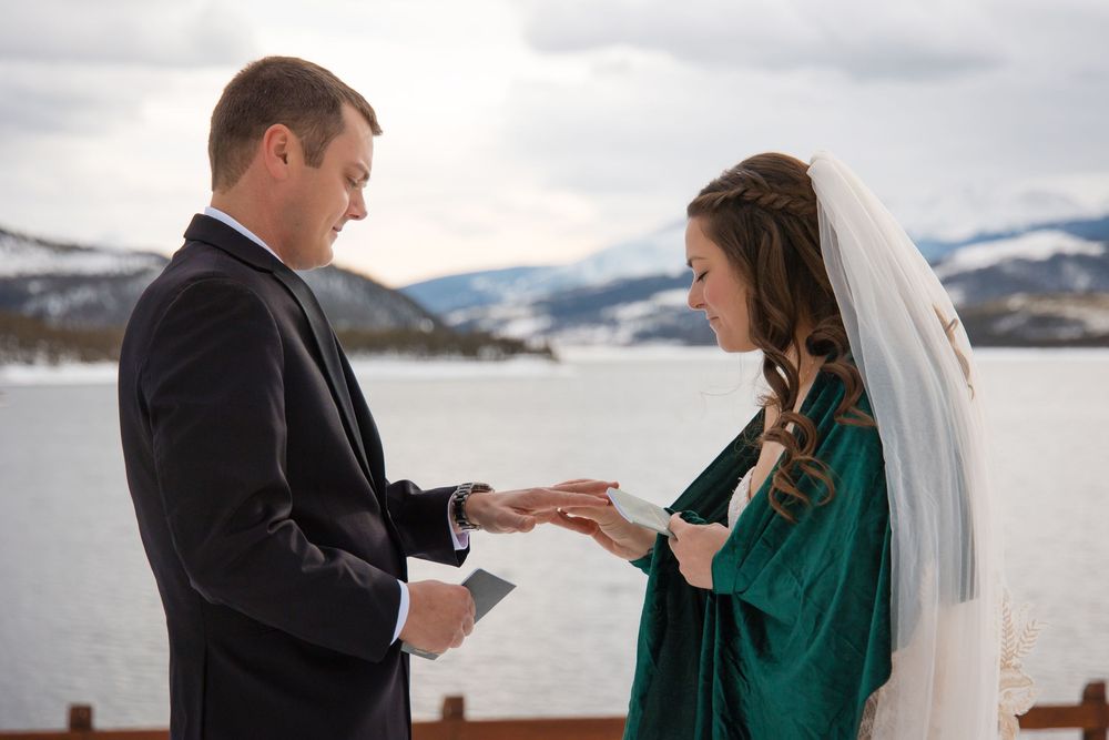 Bride in green dress and veil exchanges rings with groom in black suit during outdoor winter wedding ceremony.
