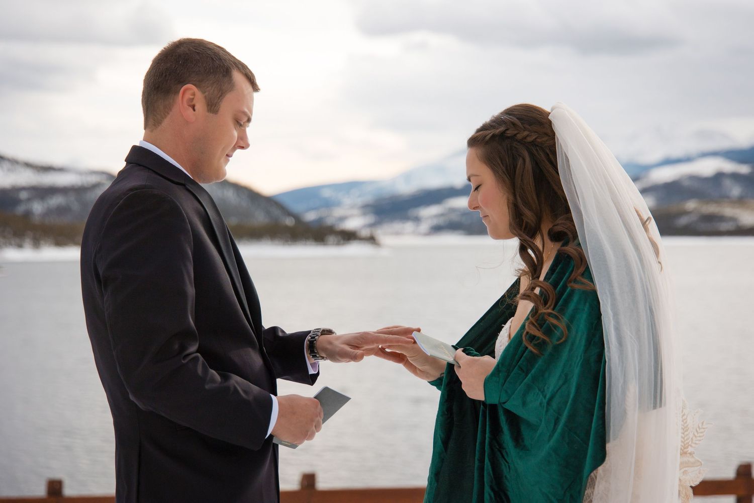 Bride in green dress and veil exchanges rings with groom in black suit during outdoor winter wedding ceremony.