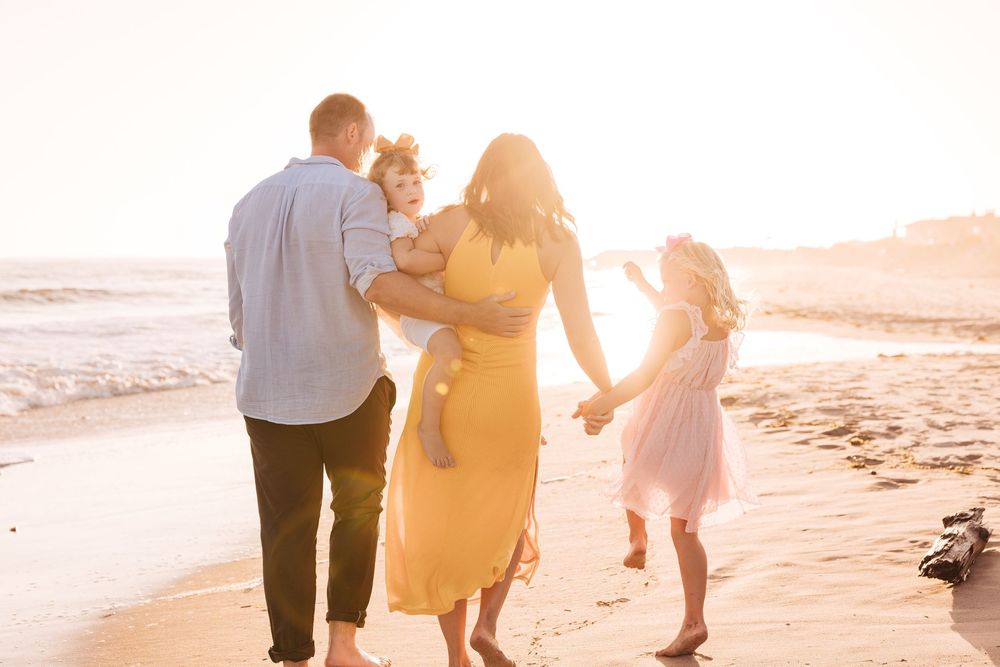 Family of four walking barefoot on a sunny beach at golden hour, parents holding children's hands near the ocean waves.