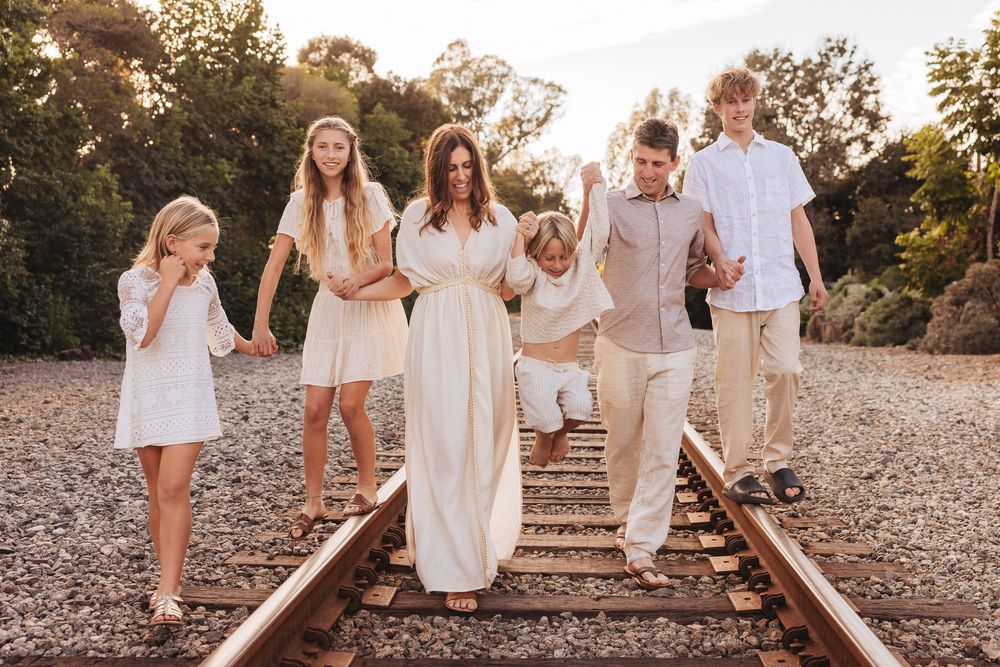 Family of six walking on railroad tracks in coordinated white and beige outfits during golden hour sunset.