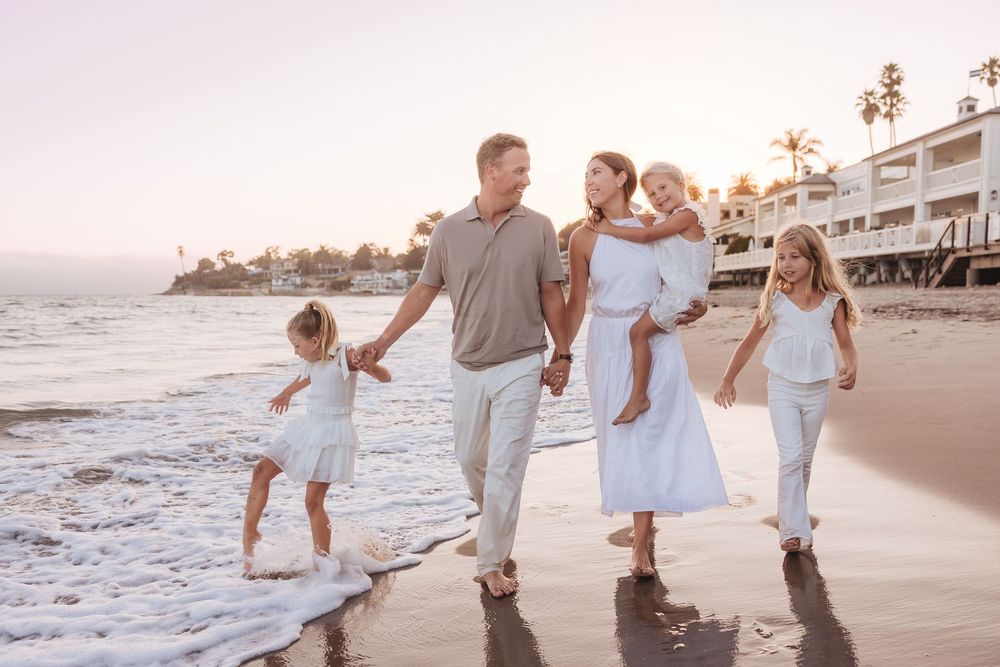 Family of five walking on a sandy beach at sunset, dressed in white and light colors, smiling and holding hands.