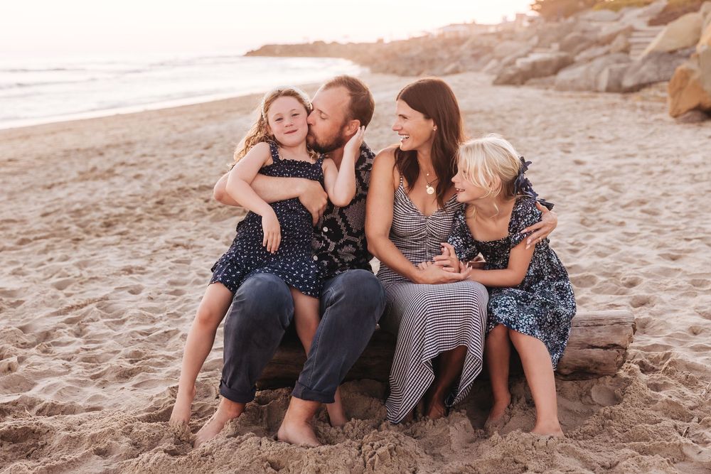 Family of four sitting on driftwood at beach during golden hour, smiling and embracing in coordinated outfits.