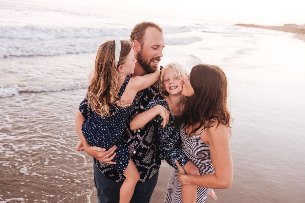 Family of four sharing joyful kisses on a sunny beach, parents holding two young daughters by the ocean waves.