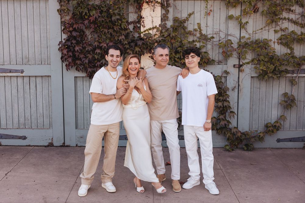 Family of four posing together in white and beige outfits in front of rustic doors with ivy backdrop.
