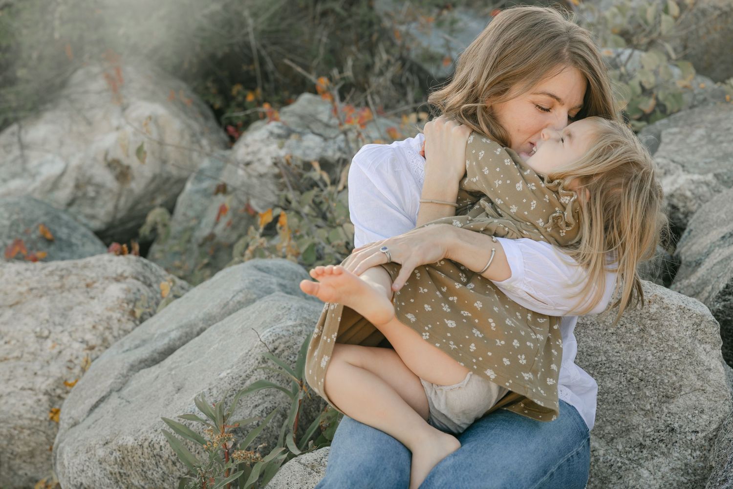 A mother and young daughter share a tender embrace while sitting on rocky terrain outdoors.
