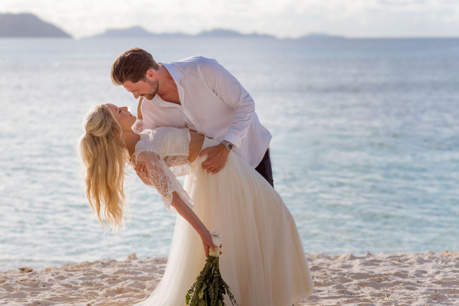 Bride and groom share a romantic kiss on a tropical beach, with turquoise ocean waters in the background.