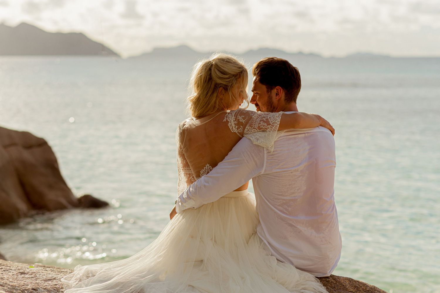 Bride and groom sitting on rocks by turquoise ocean, sharing a romantic embrace in wedding attire at sunset.