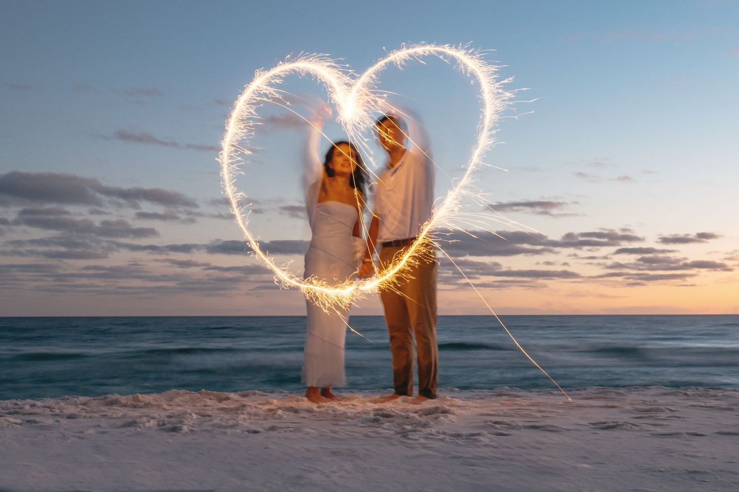 Couple draws heart shape with sparklers on Fort Walton beach during sunset creating romantic silhouette against ocean. MJ Photographs
