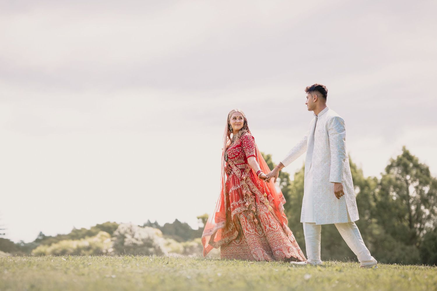 Couple in traditional Indian wedding attire walking hand in hand on green grass, bride in red lehenga, groom in white sherwani.