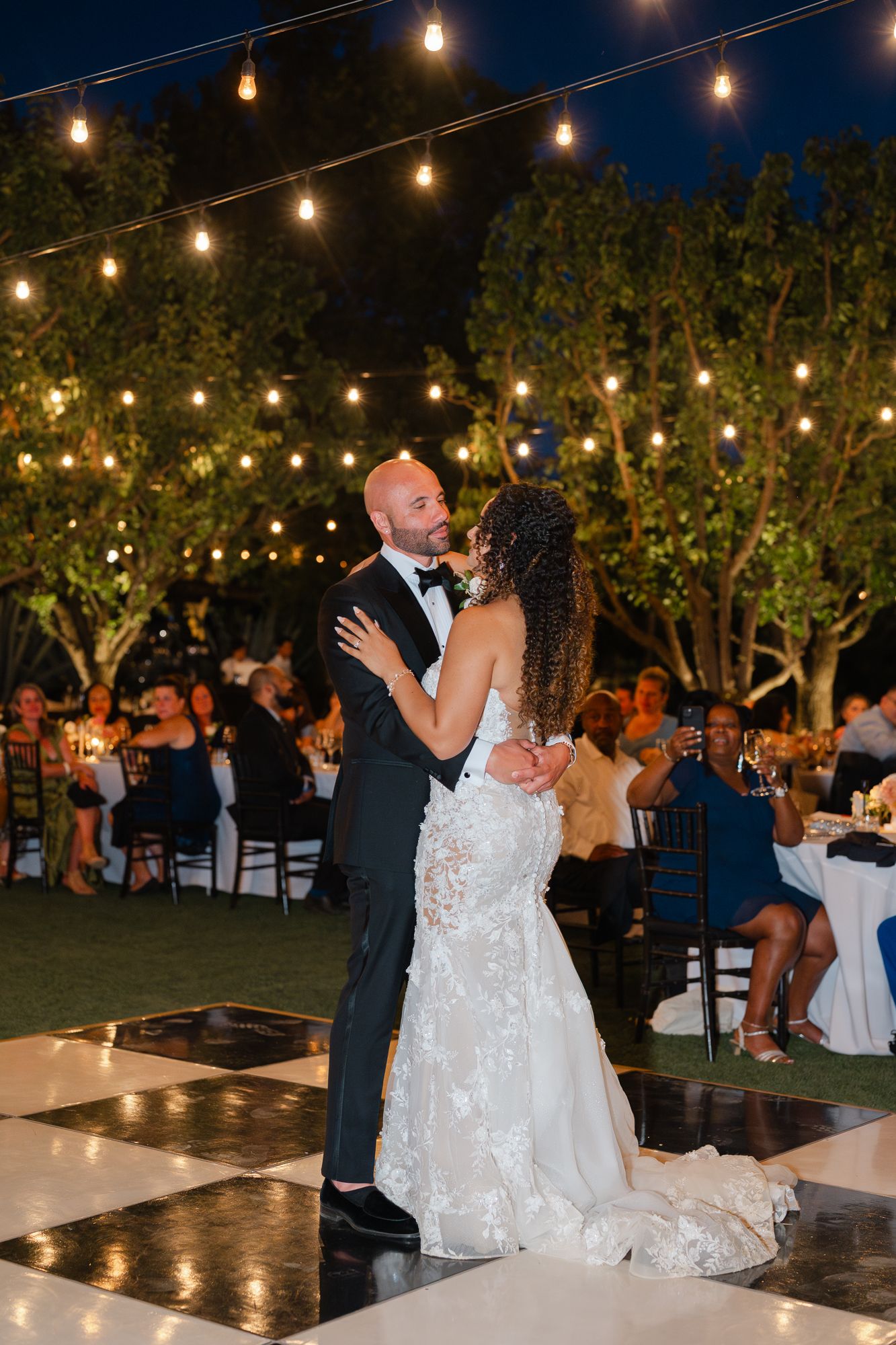Couple shares first dance on checkered floor under string lights at outdoor evening wedding reception.