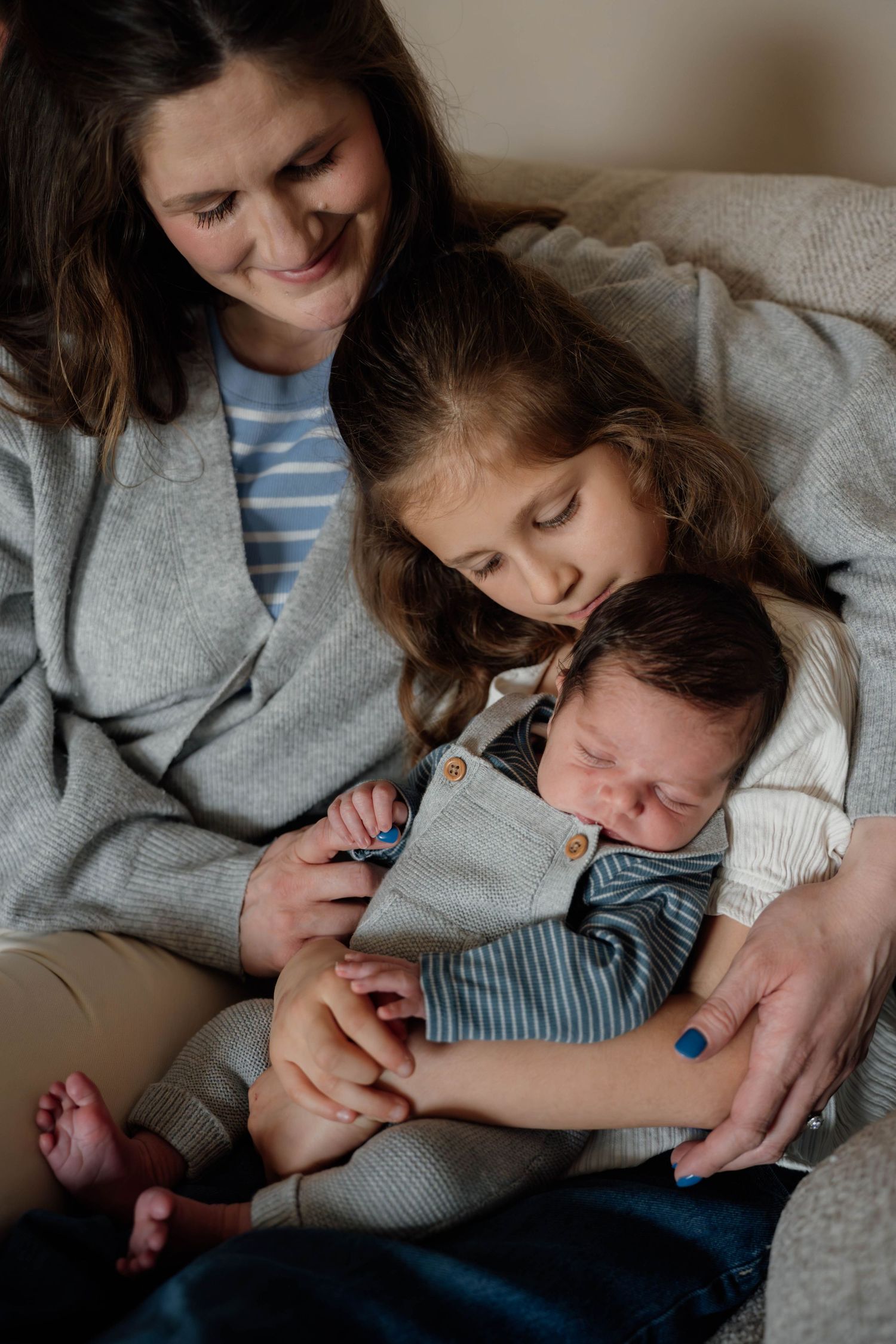 A smiling mother sits on a couch with her young daughter kissing a sleeping newborn baby wrapped in a gray blanket.
