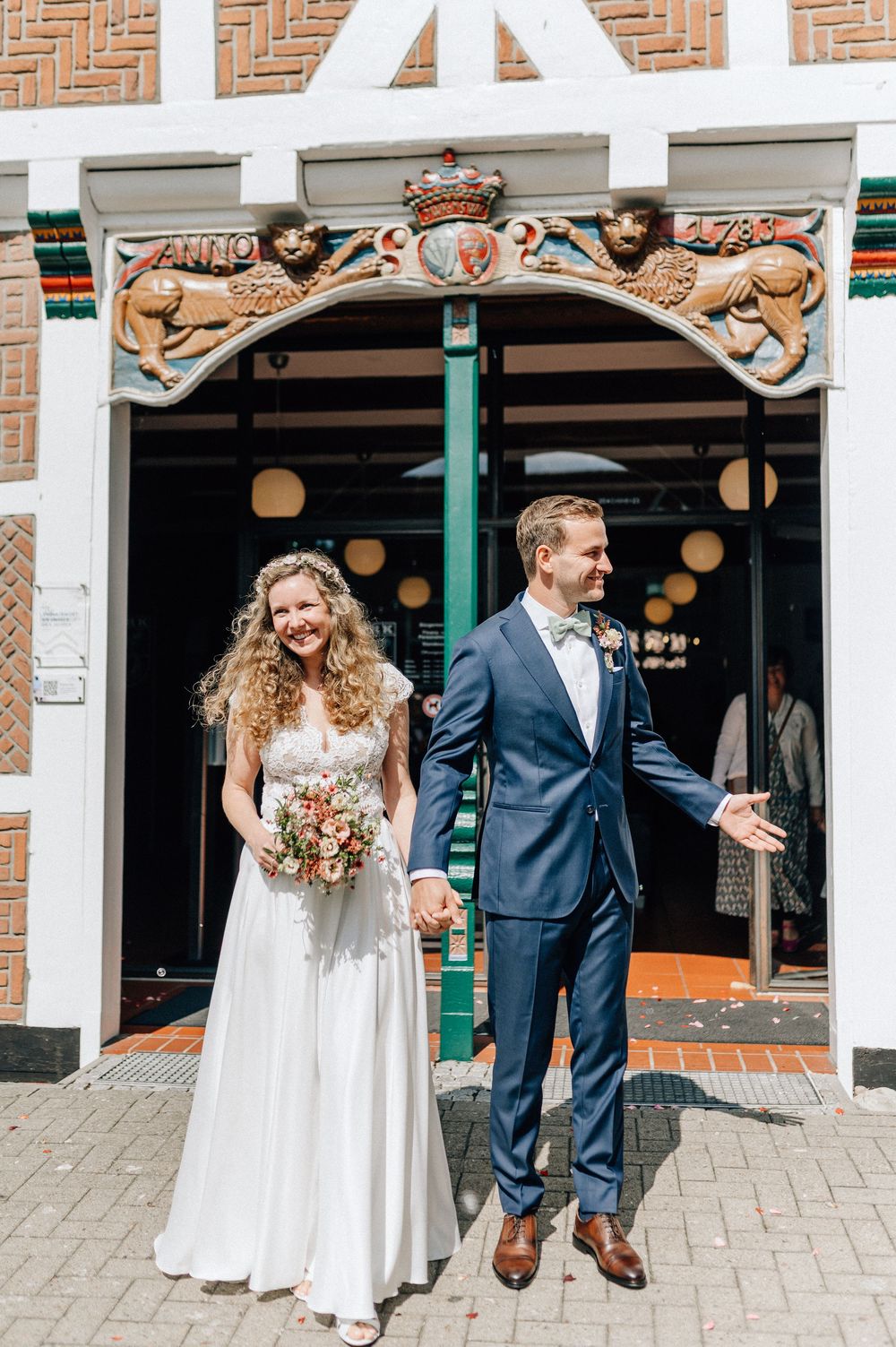 Bride and groom holding hands outside a historic building, bride in white gown with bouquet, groom in navy suit.