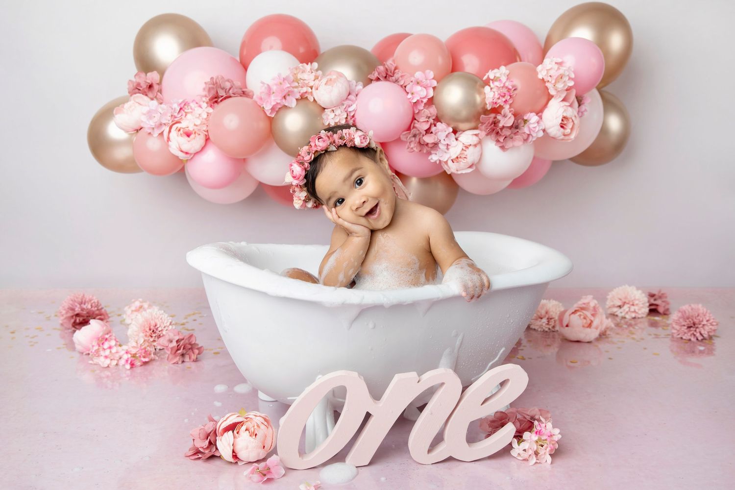 Baby girl in white bathtub with pink balloons, flowers, and “one” sign celebrating her first birthday.