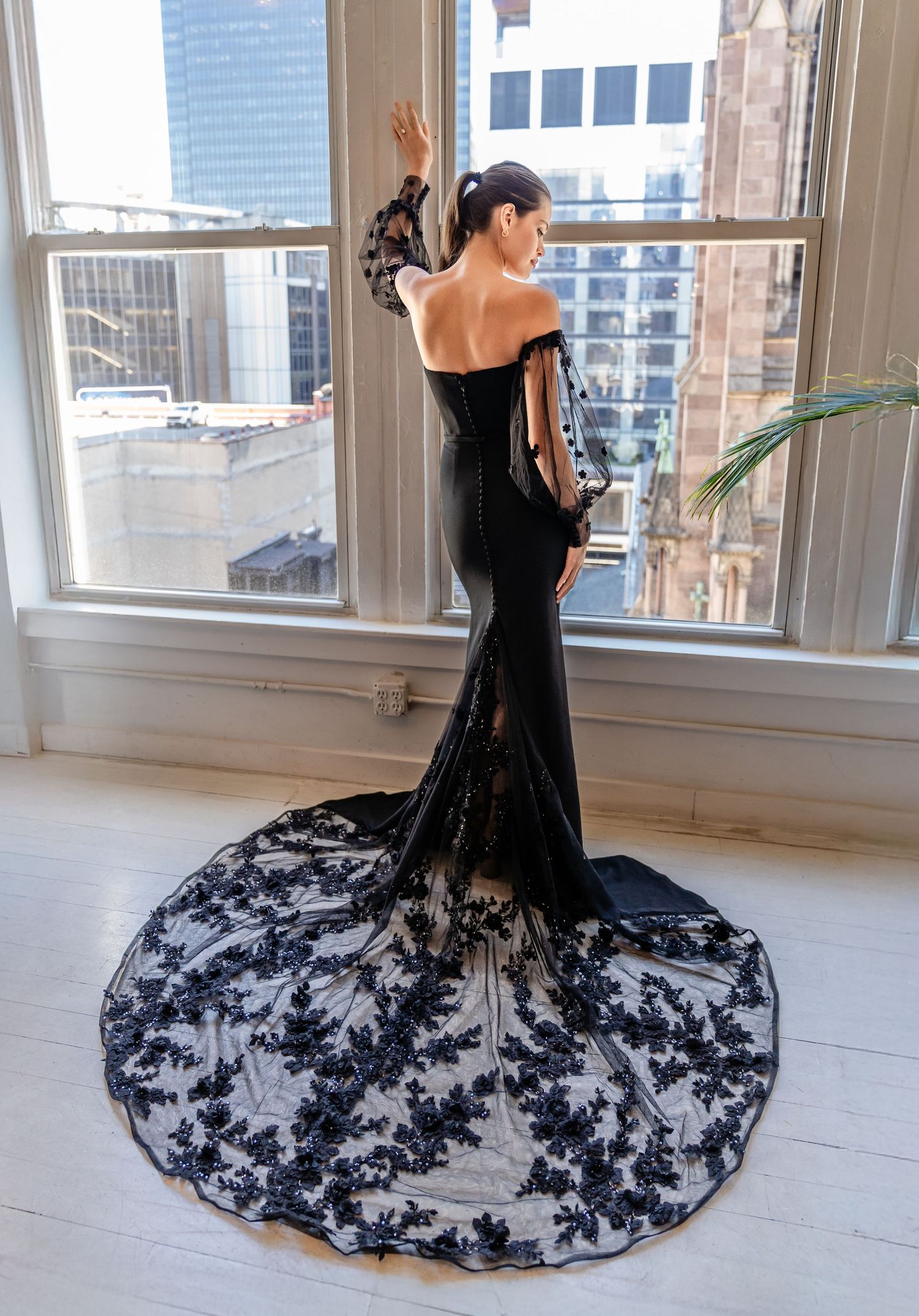 Black lace mermaid gown with long sleeves and dramatic train, worn by model posing at window in urban setting.