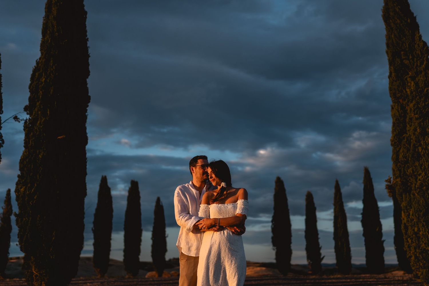 Couple embracing at sunset on a Tuscan cypress-lined path with dramatic cloudy sky in Italy.