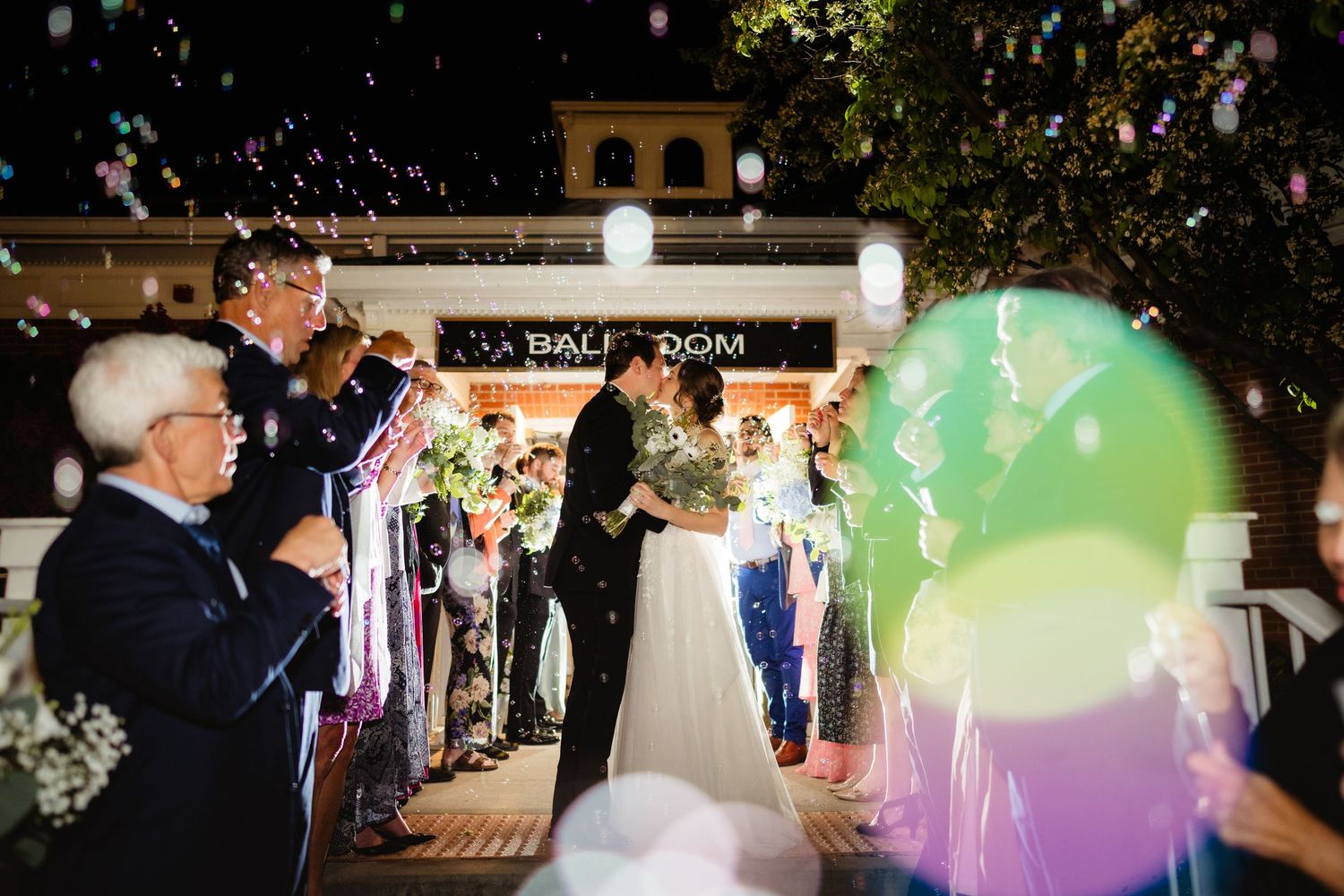 Wedding couple exits ballroom at night amid colorful confetti and balloons, surrounded by cheering guests.