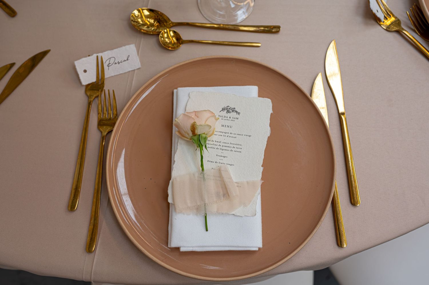 Cadre de mariage élégant avec couverts en or, plaque de chargeur blush, serviette blanche et une seule rose sur une nappe neutre.