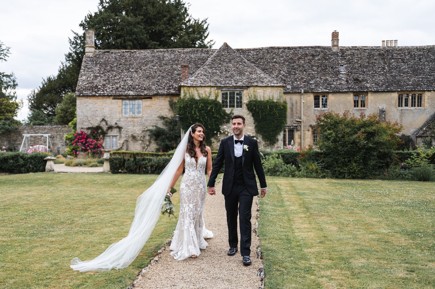 Bride with long veil and groom in black tie walking past a beautiful Cotswold stone manor house at Caswell House Wedding Venue