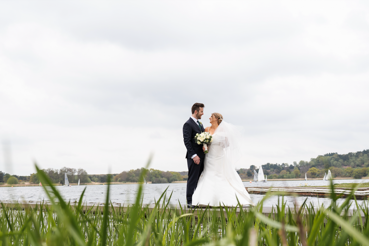 Bride and groom stand close together on a dock with a serene lake backdrop.