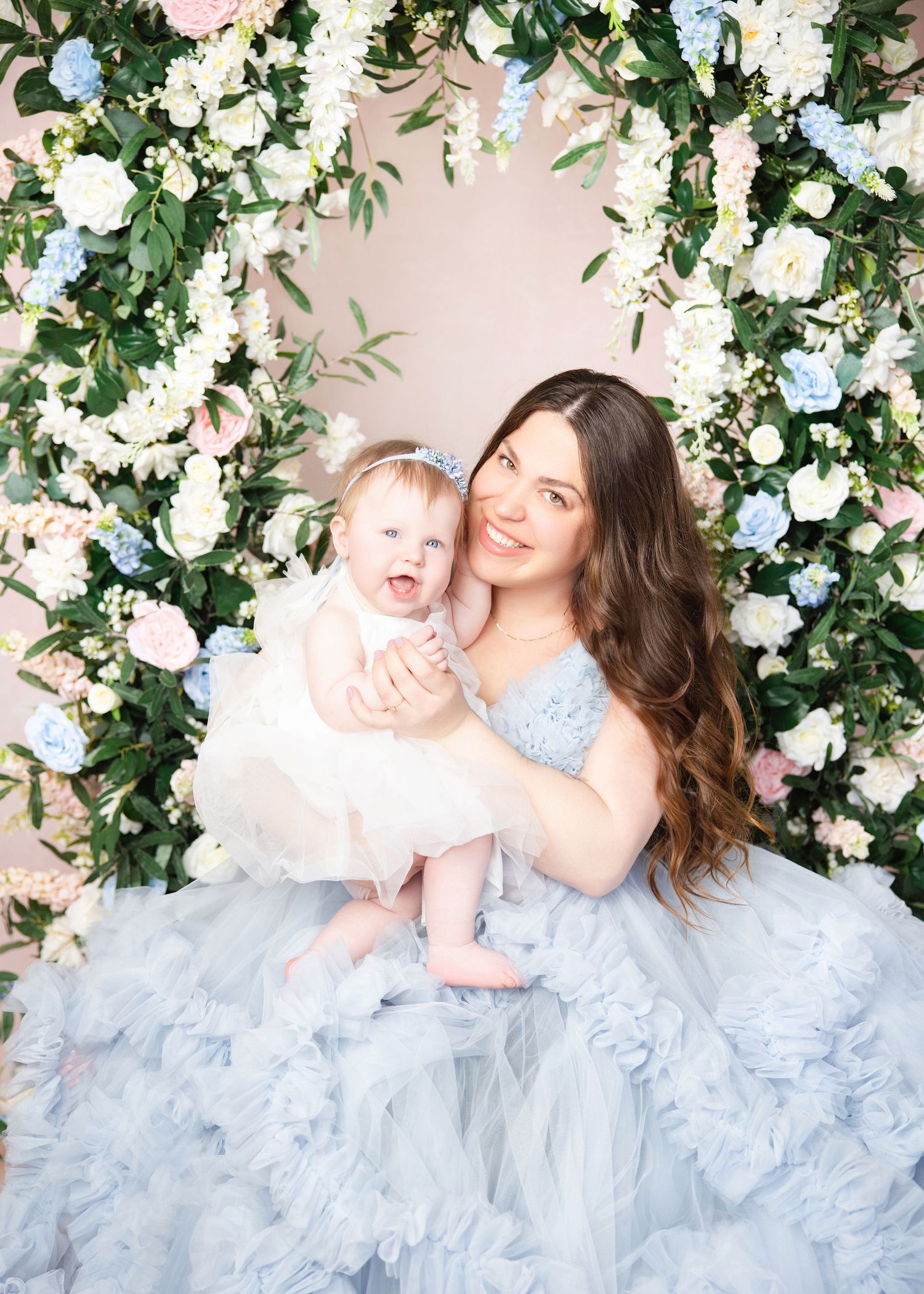 A smiling woman in a blue tulle gown holds a happy baby girl, both posed before a floral arch of white and pink blooms.