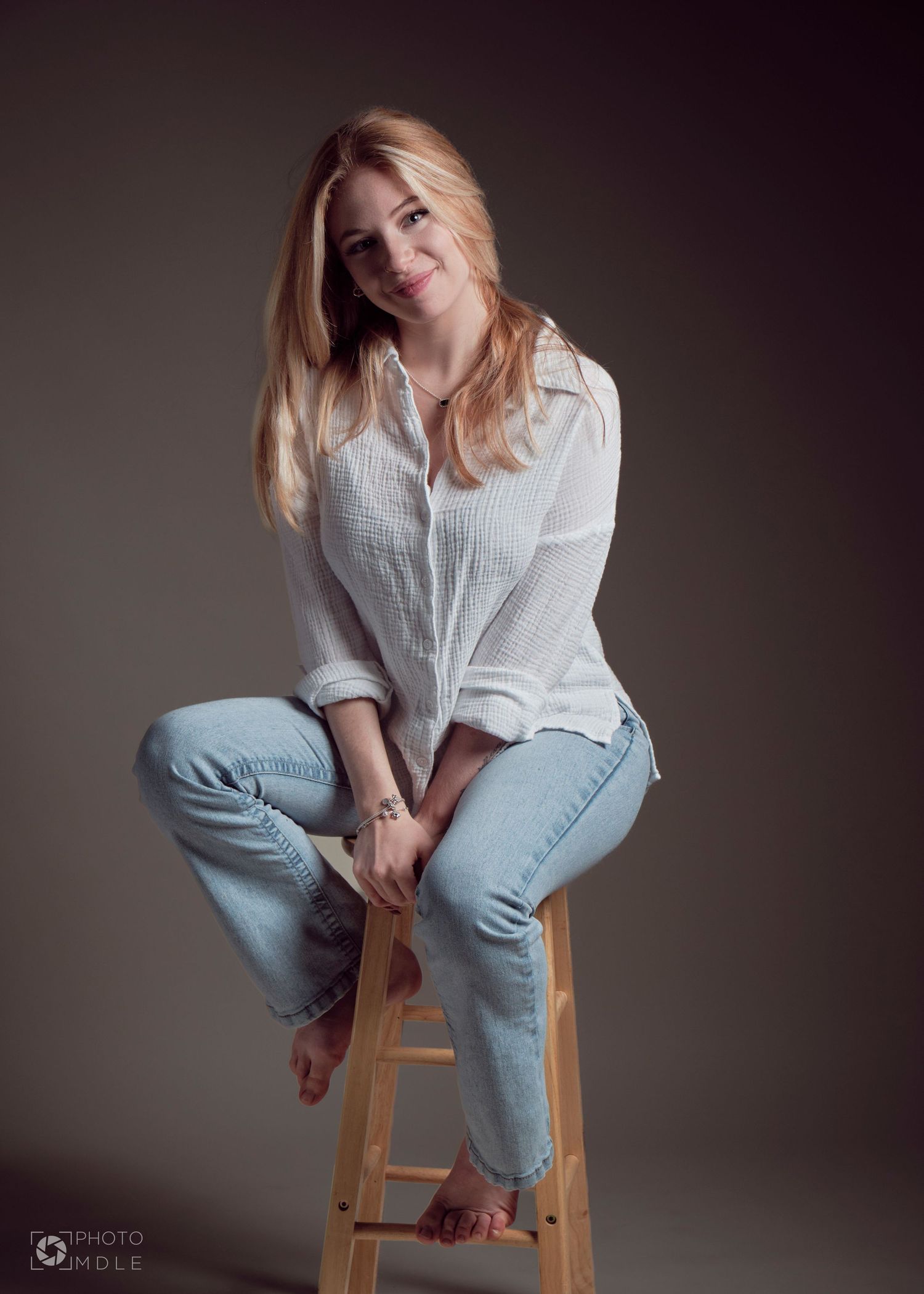 Une jeune femme assise sur un tabouret, pieds nus, regarde la caméra avec un sourire paisible.