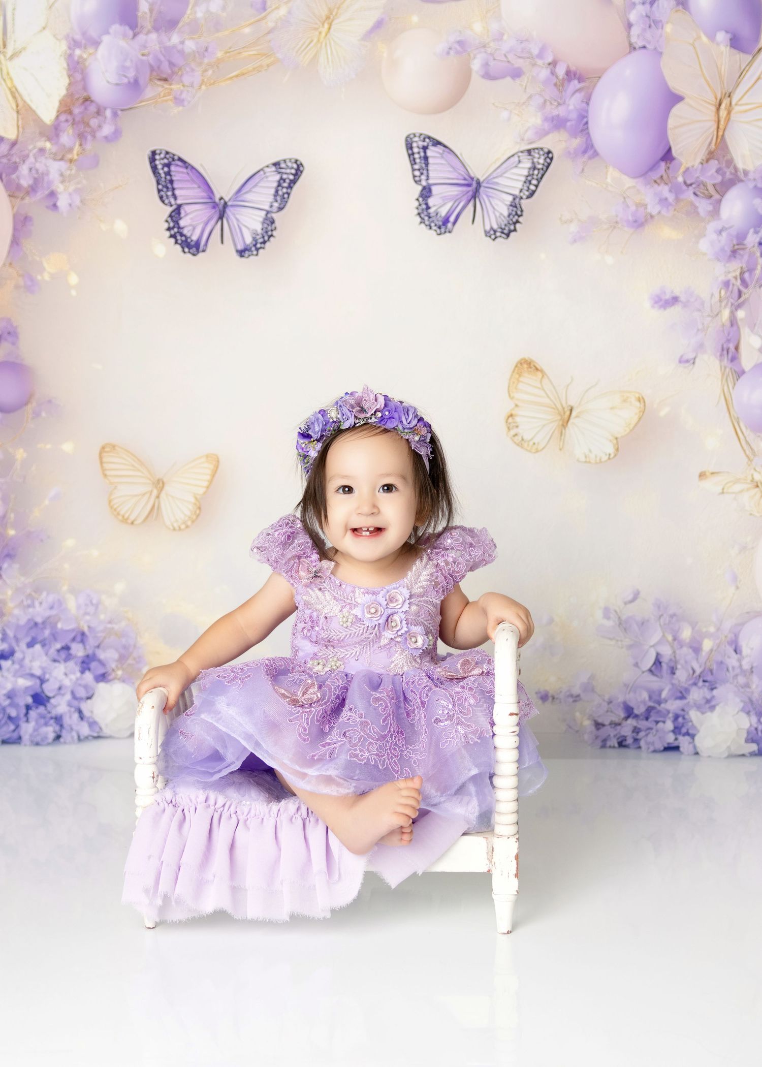 Smiling toddler girl in purple lace dress sits on white bench with butterfly and balloon backdrop.