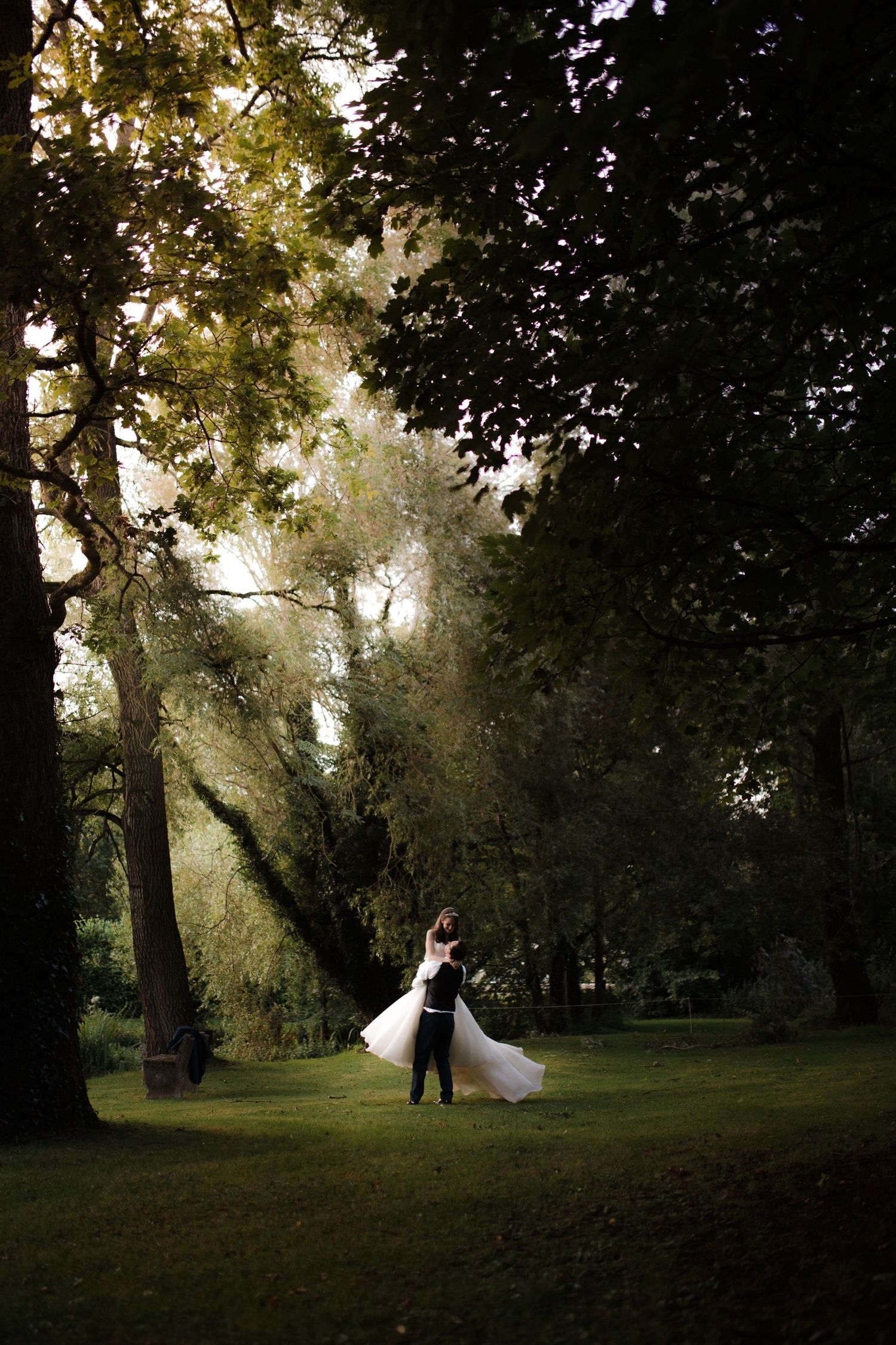 Bride and groom embracing in a lush green park, sunlight filtering through tall trees on their wedding day.