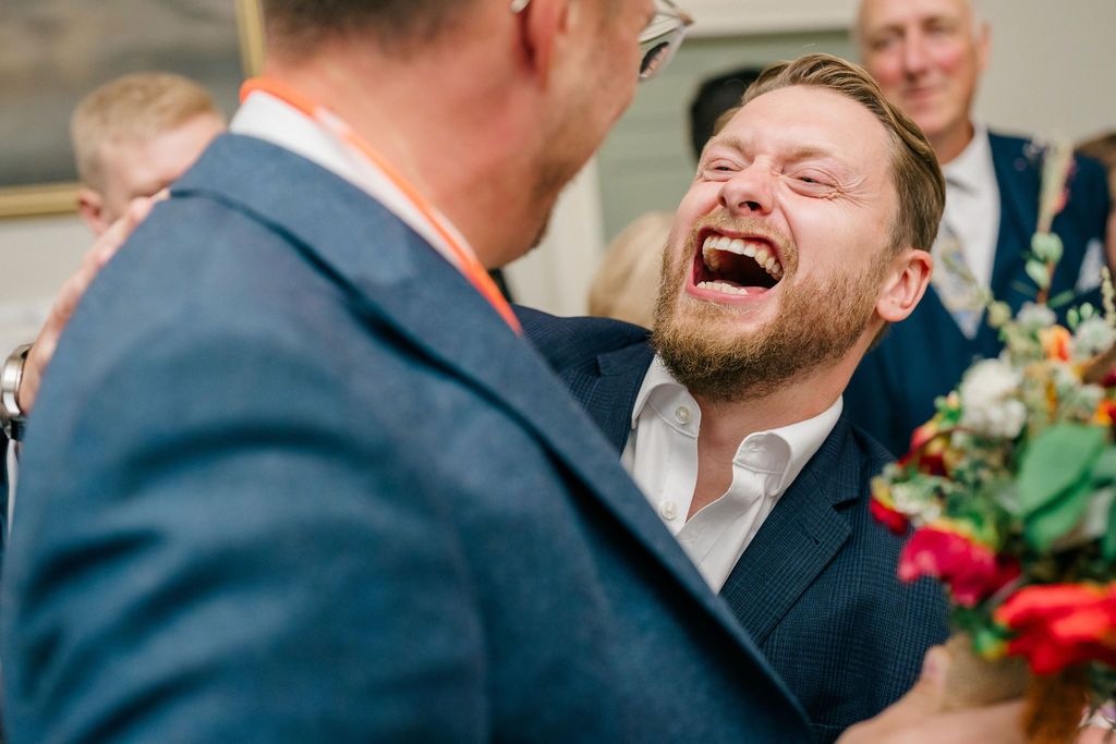 Two men in navy suits laughing joyfully at a wedding celebration, one holding a colorful bouquet.