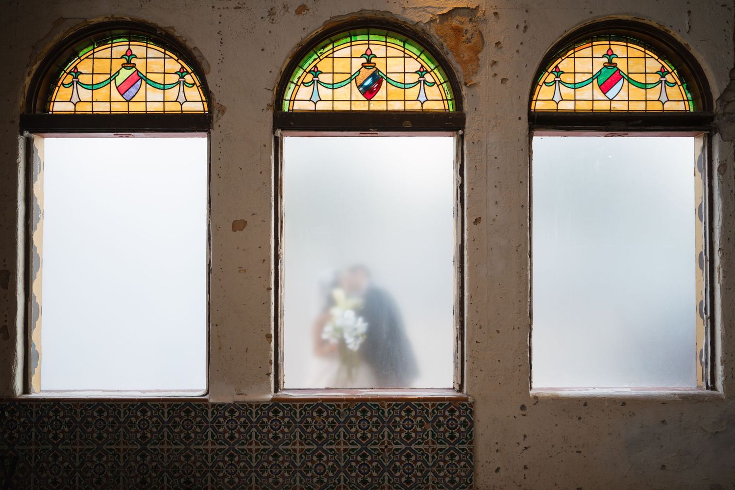 Couple kissing behind frosted glass windows with colorful stained glass arches in a rustic vintage building.