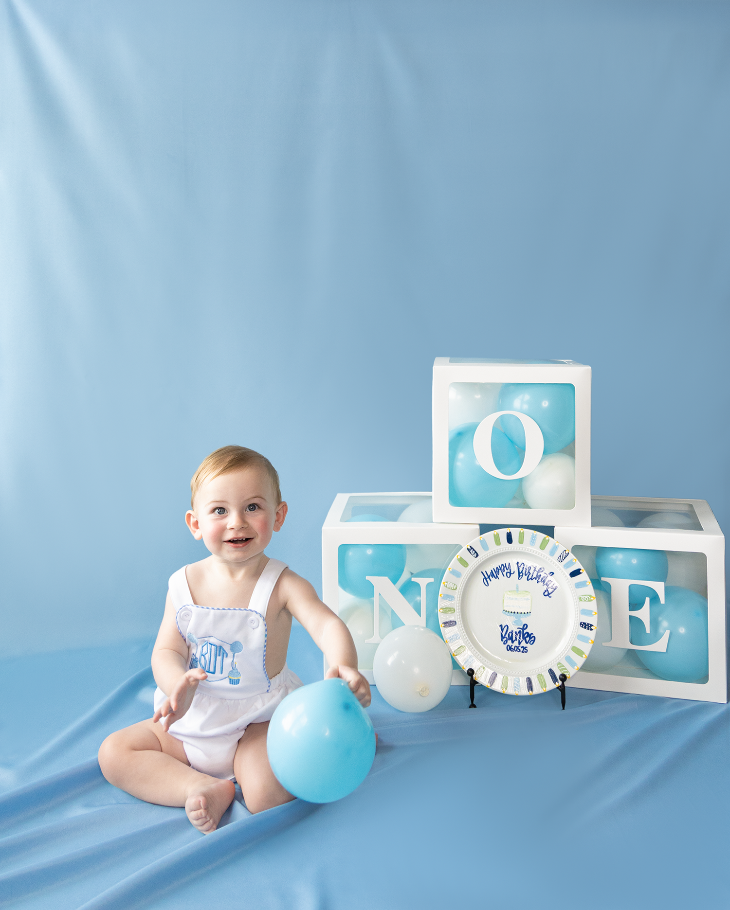 Smiling baby boy sits with blue balloons beside letter blocks spelling ONE on a blue backdrop for first birthday.