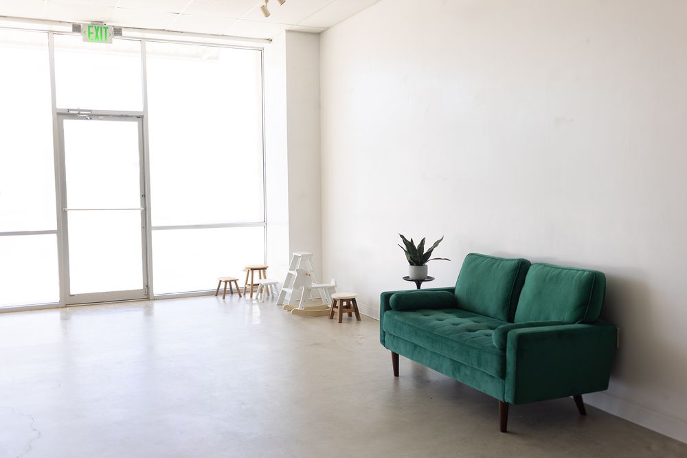 Minimalist studio space with green velvet sofa, white walls, concrete floors, and natural light from large windows.