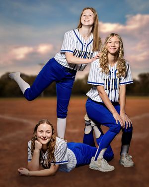 Three girls in blue and white Twins softball uniforms pose together on a baseball field backdrop.