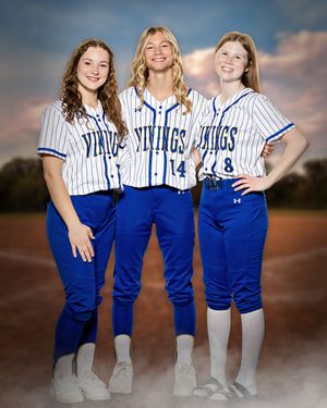 Three girls in Vikings softball uniforms smiling together on baseball field backdrop.