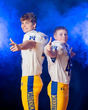 Two Minneota Vikings football players pose back-to-back in blue and gold uniforms against a blue smoke background.
