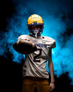 Minneota Vikings #2 football player in gold helmet extending football toward camera in blue smoke.