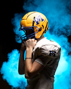 Football player in gold Minneota helmet holds facemask dramatically against blue smoke background.