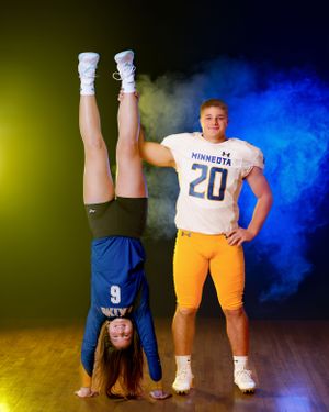 Gymnast does a handstand beside Minneota football player number 20 against colorful smoke backdrop.