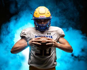 Minneota Vikings football player in gold helmet holding football in dramatic blue smoke.