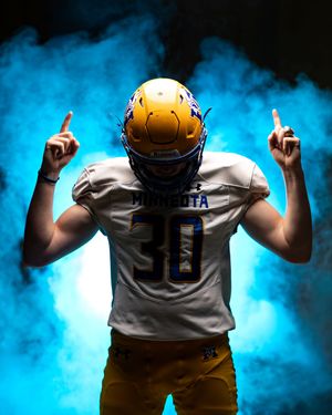 Minneota football player number 30 in gold helmet points upward in blue smoky background.