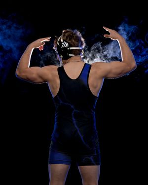 Wrestler in dark singlet and headgear flexes both arms against a dramatic blue smoke background.