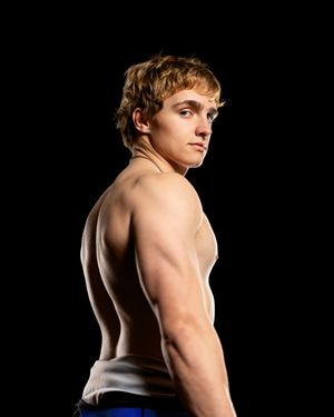 Athletic young male wrestler poses shirtless in profile against a dark background showcasing muscular build.