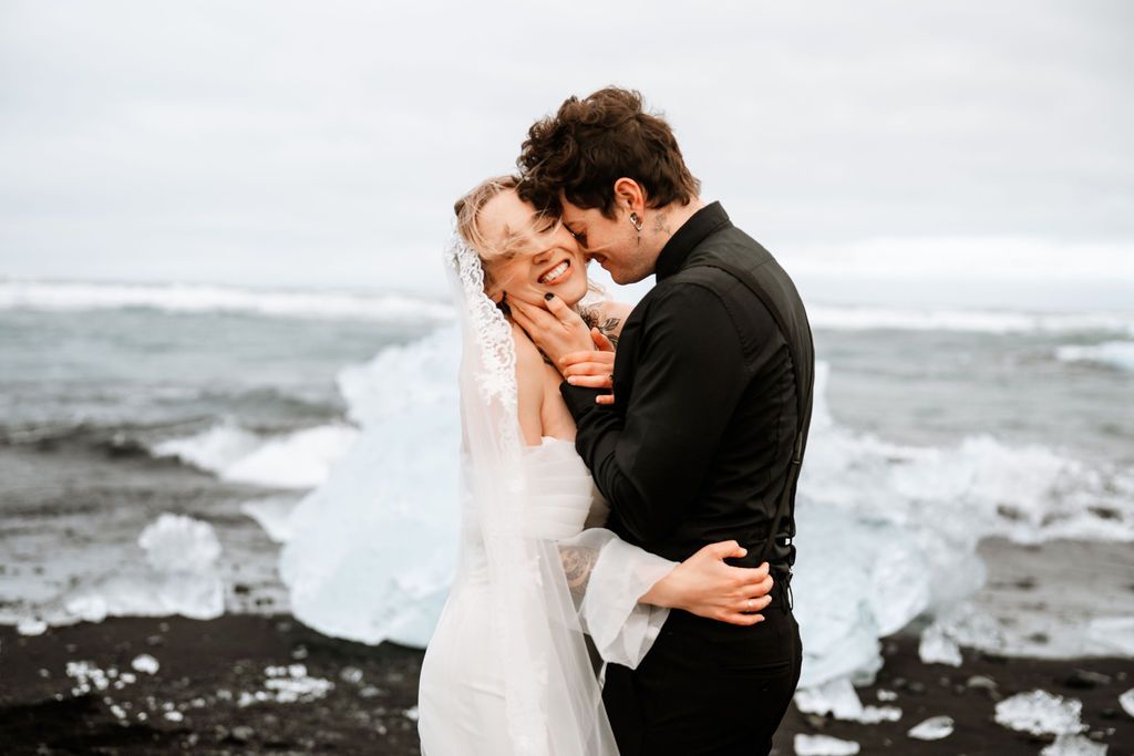 Bride and groom share romantic moment near glacial lagoon in Iceland, bride holding red bouquet in white gown.