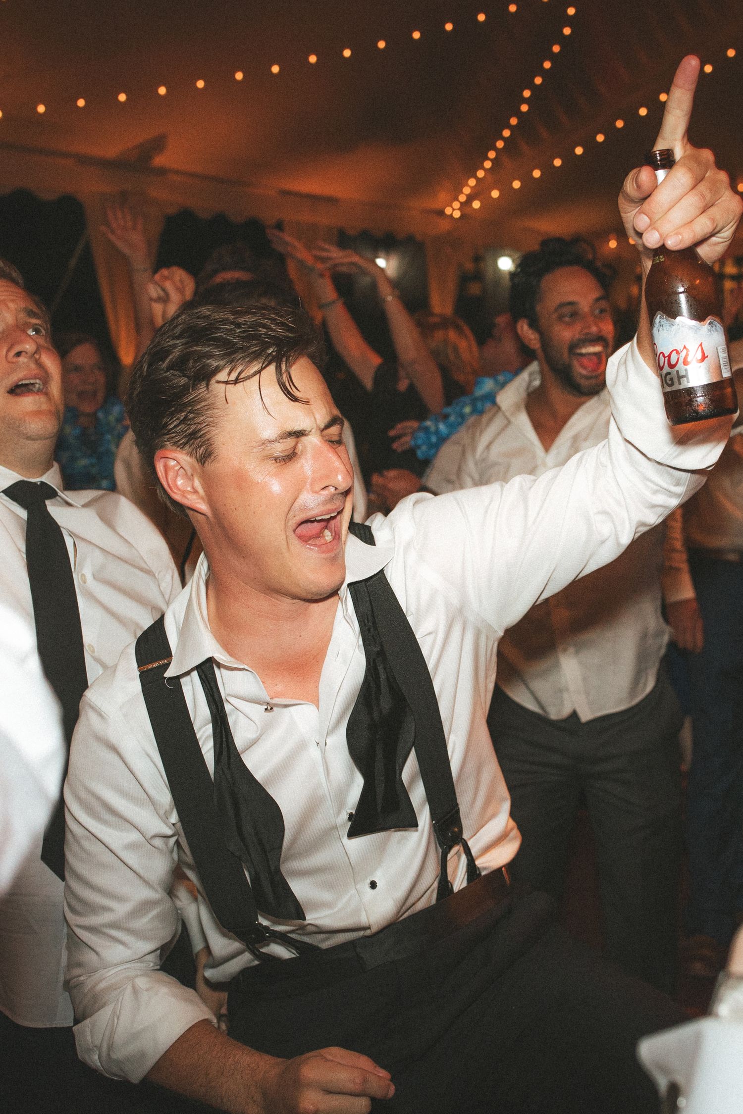 Man in suspenders raises Coors beer bottle at lively wedding reception party celebration.