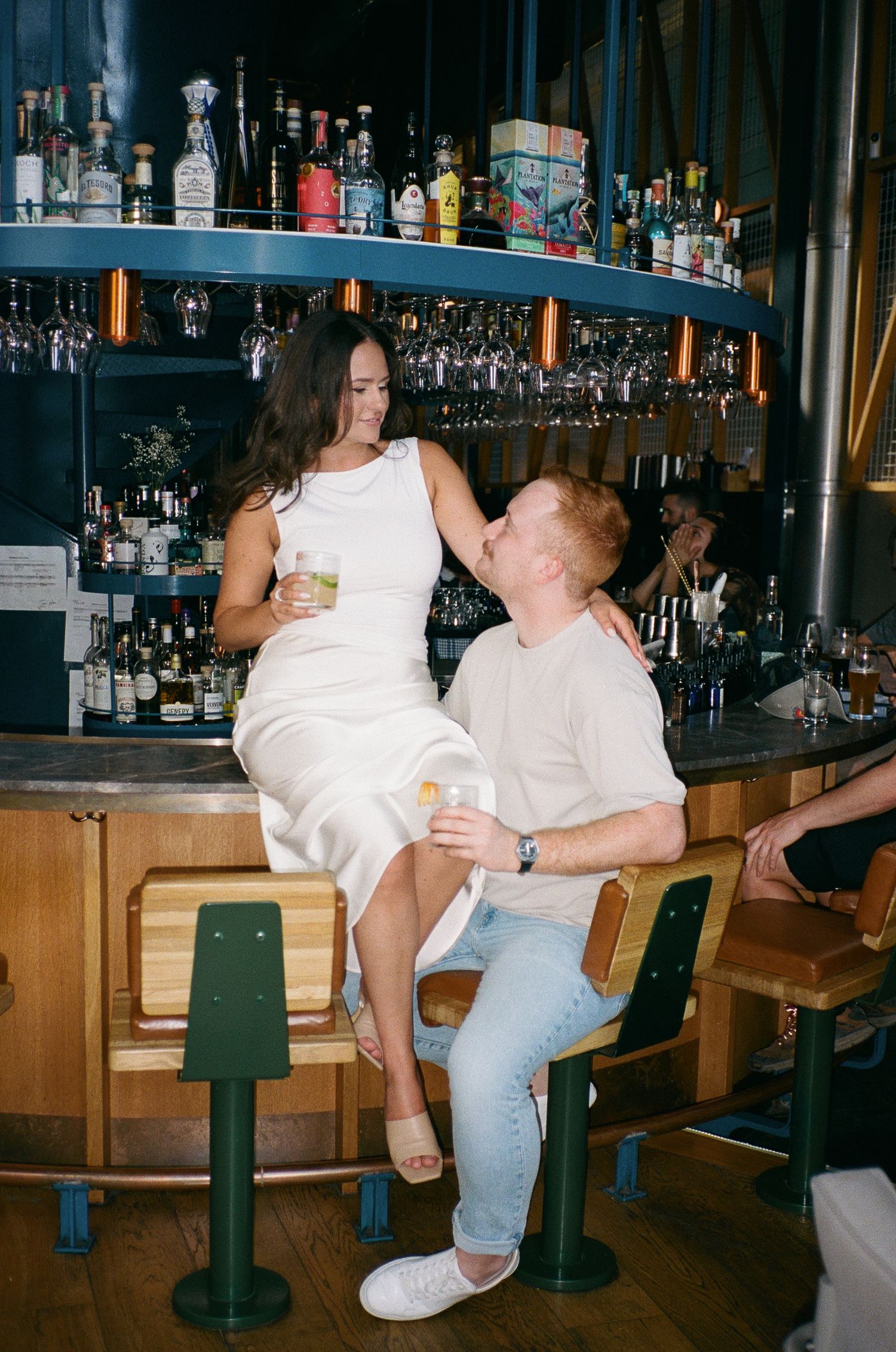 Couple sharing a romantic moment at a bar, fiancé in white dress seated on counter, man in beige shirt at bar. Aliza Dworkind photoshoot
