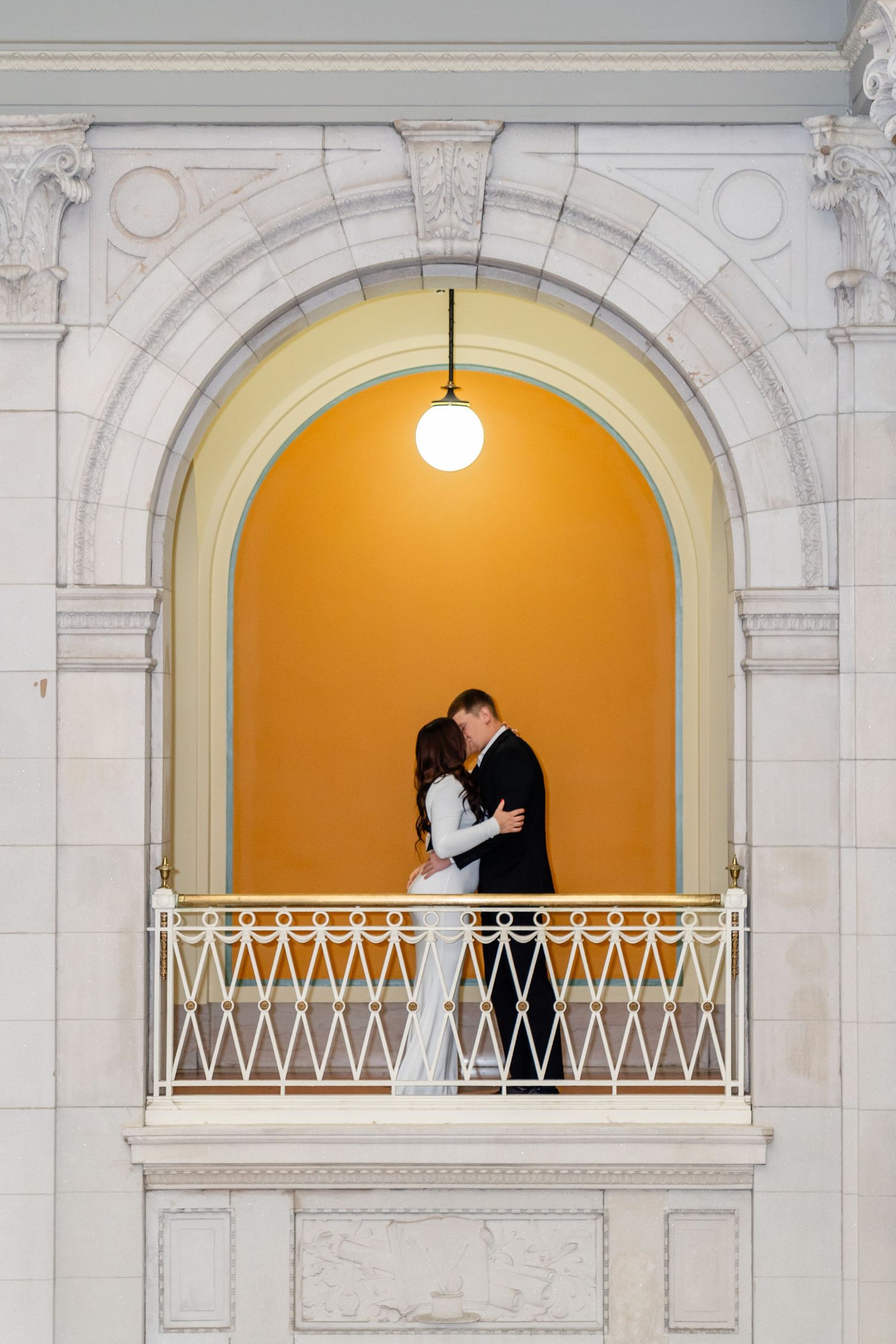 Couple sharing a romantic kiss in an elegant marble building, man in black suit, woman in white fitted gown.
