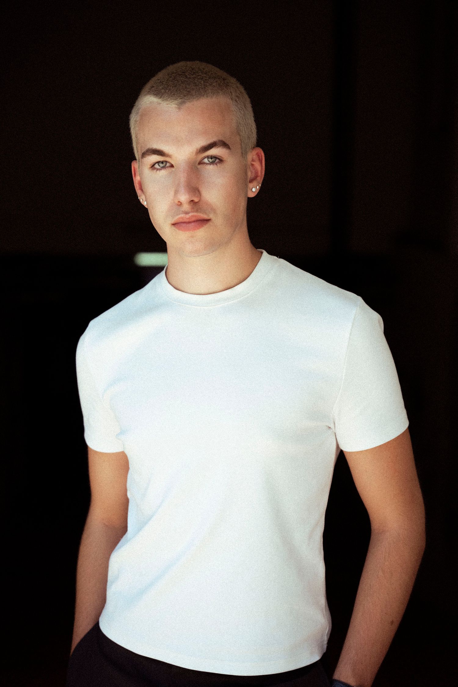 Young man with buzzed blonde hair wearing a white t-shirt posing against a dark background.