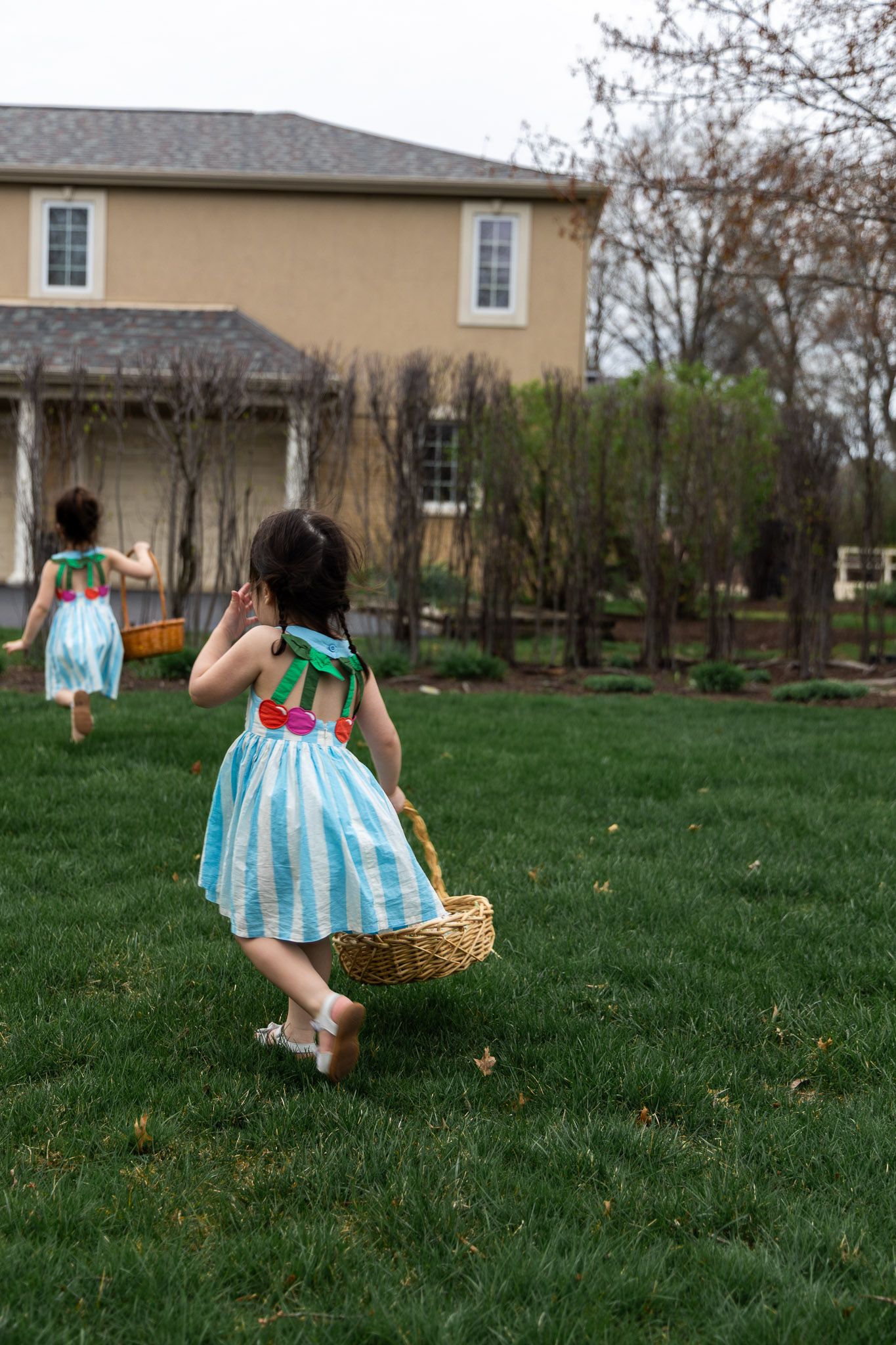 Two young girls in colorful striped dresses carrying baskets run across a green lawn during an Easter egg hunt.