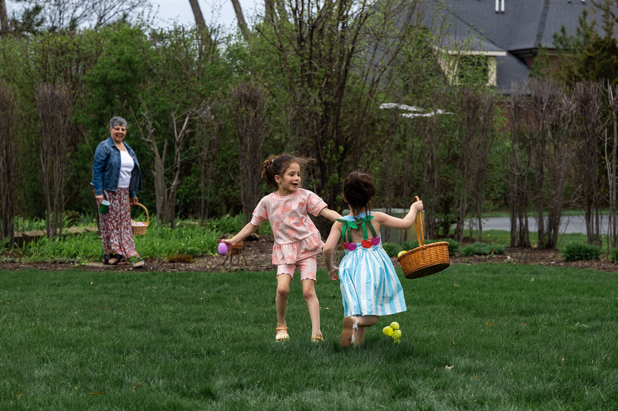 Two young girls hold hands playing in a green backyard while a woman watches during an Easter egg hunt.