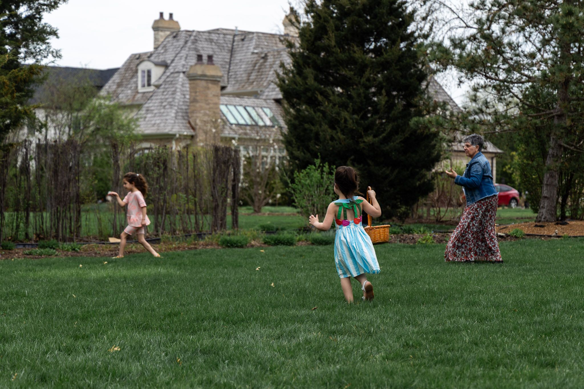 Two children and an adult playing outdoors on a green lawn with a large stone manor house in the background.