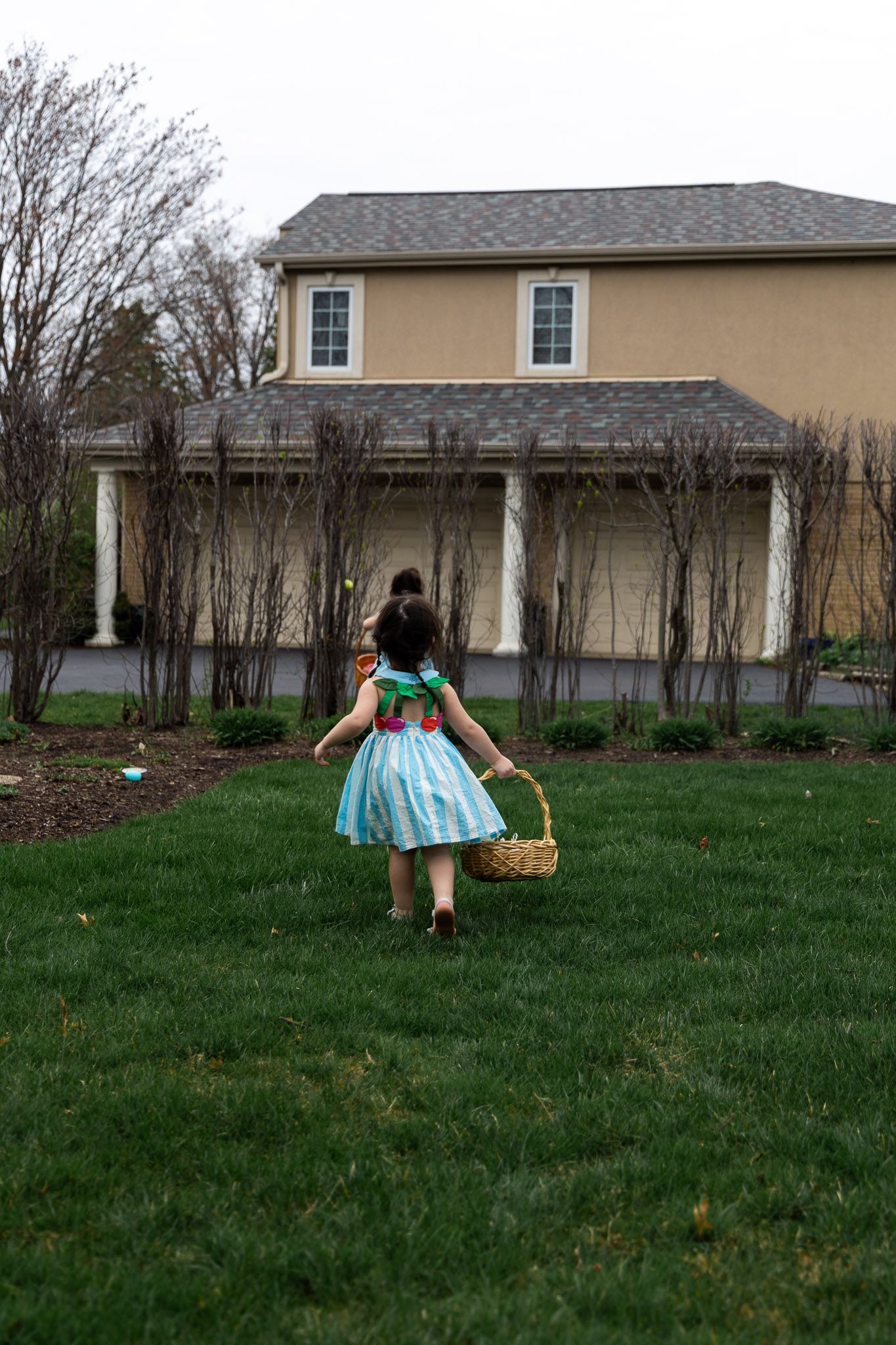 Young girl in blue dress carrying Easter basket on green lawn in front of large two-story suburban home.